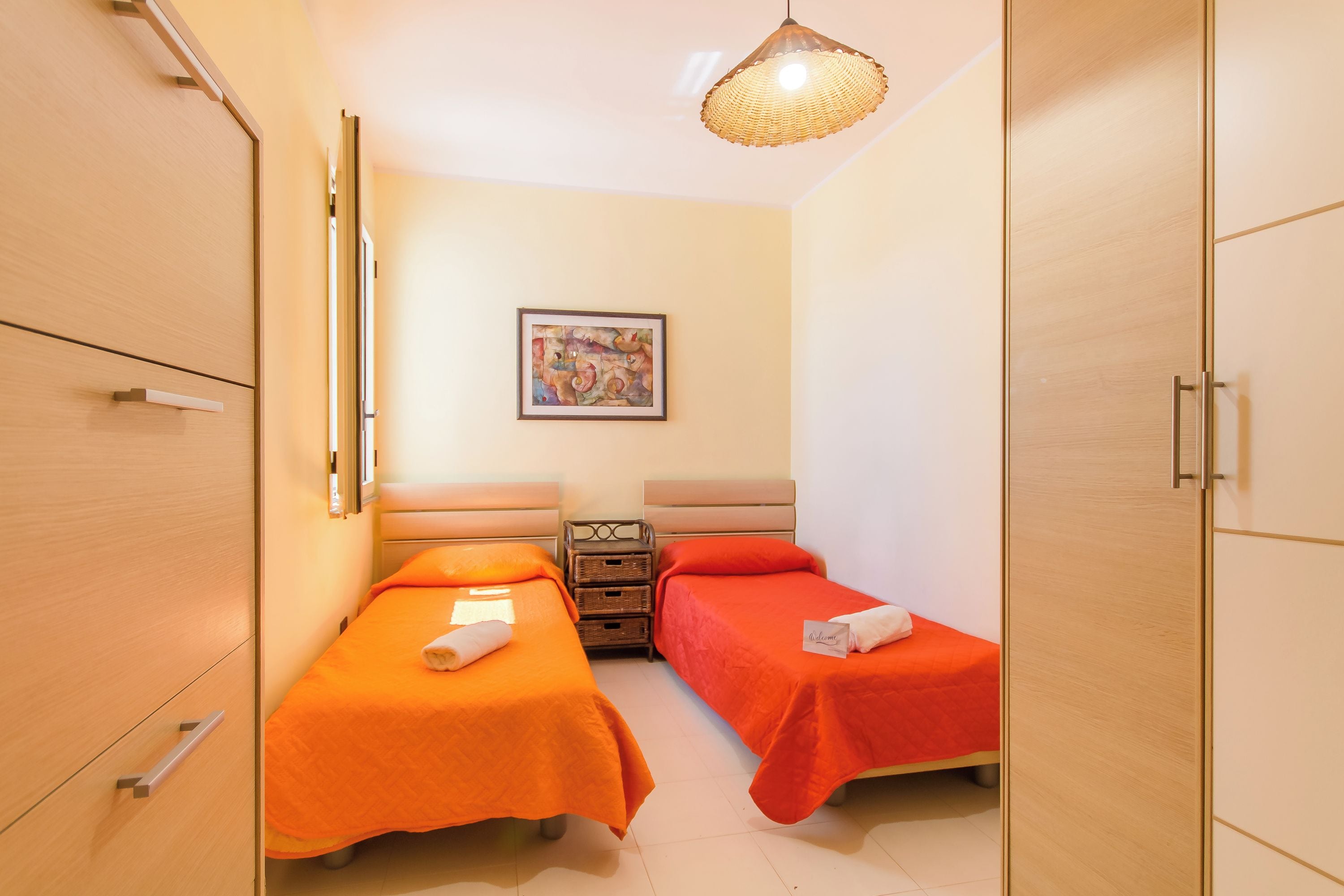 A hotel room with two beds, a wooden wardrobe, a wicker lamp, and a painting on the wall.