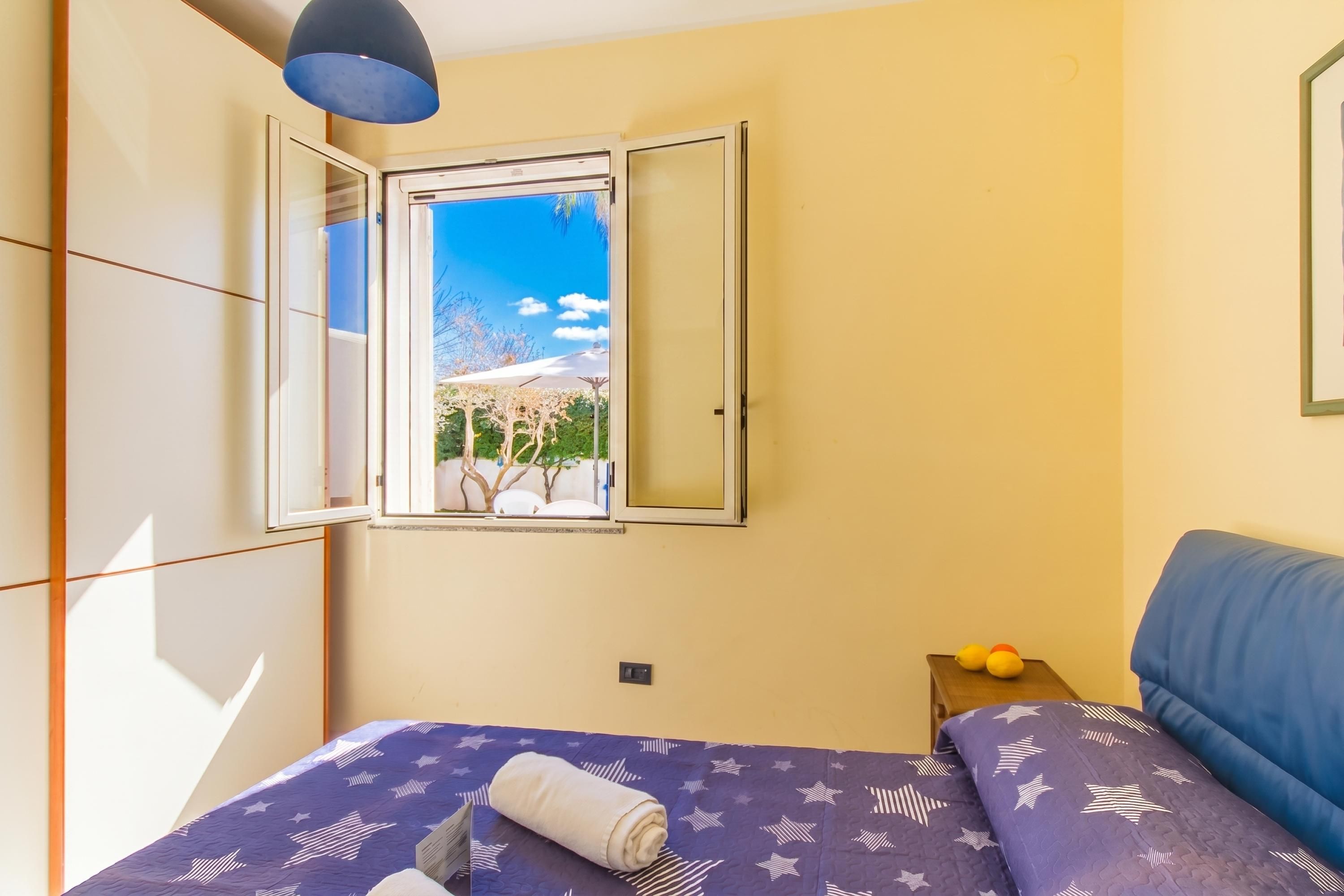 A bedroom with a bed, a window with a view, a bedside table with two yellow objects, and a blue headboard.