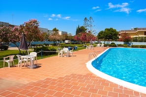 A swimming pool with a tiled deck, surrounded by white chairs and tables, and blooming pink trees. - Villetta Relax with pool in residence (Melendugno)