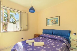 A bedroom with a bed, a nightstand, a lamp, a painting, and a window with a view of palm trees. - Villetta Relax with pool in residence (Melendugno)