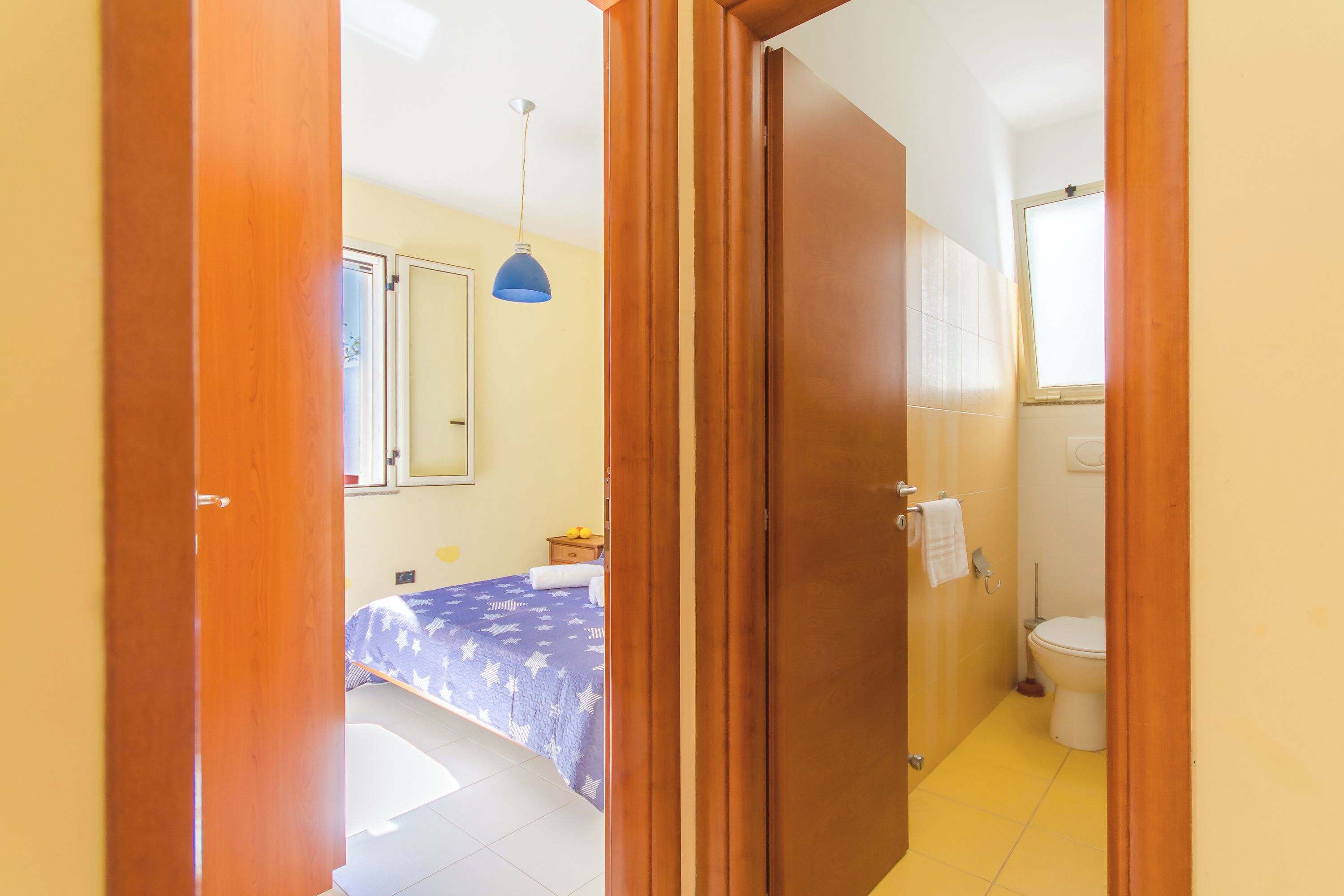 Un dormitorio con cama, mesita de noche, lámpara colgante azul y un baño con inodoro y lavabo.