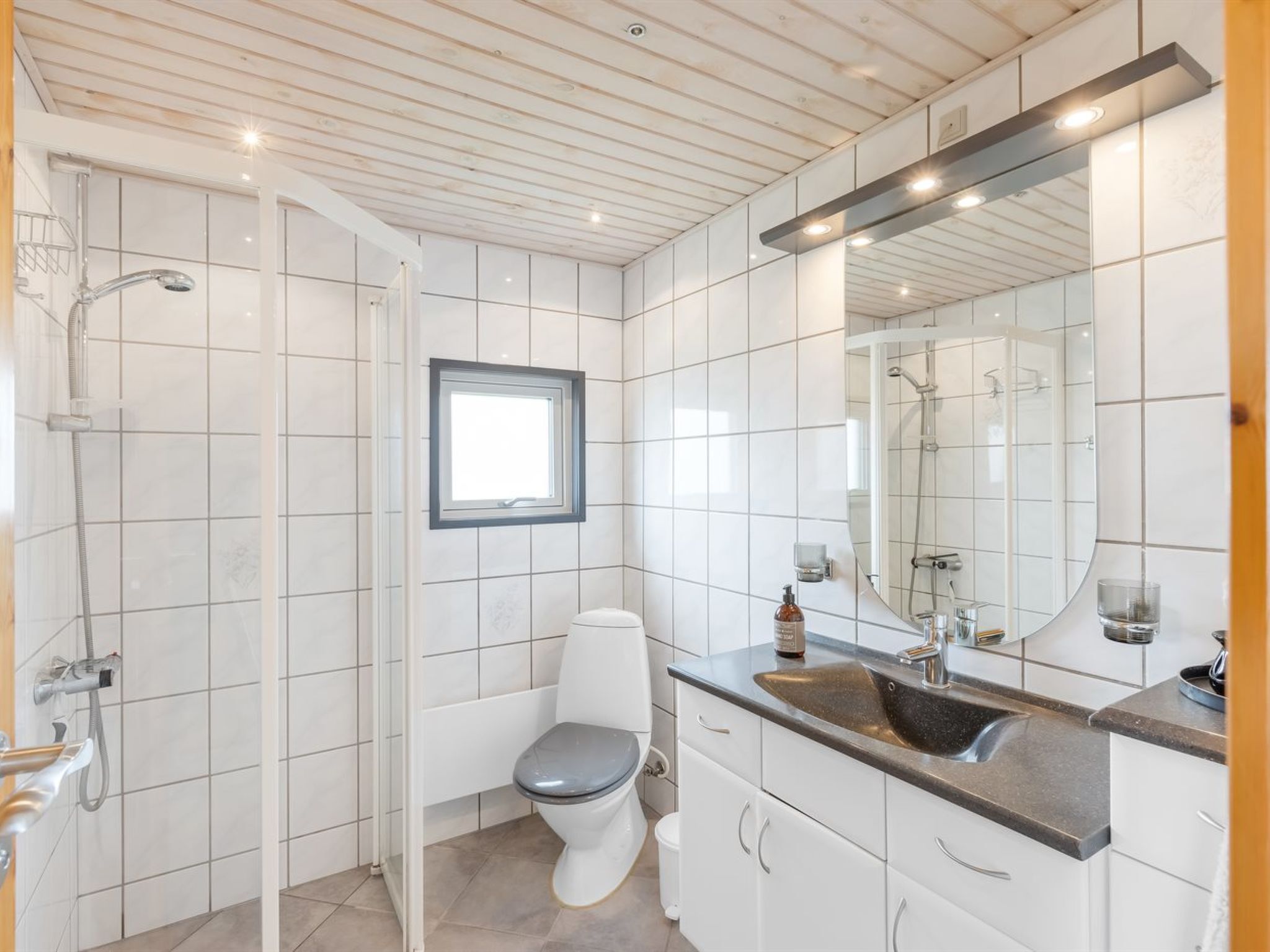 Ein modernes Badezimmer mit weißen Fliesen, Toilette, Dusche, Waschbecken mit Spiegel und einem Fenster.