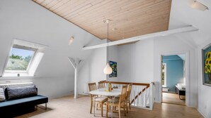 Een ruime kamer met een hoge plafond, een eethoek met houten stoelen en een witte tafel, een zwarte bank en een groot raam dat zorgt voor natuurlijk licht.