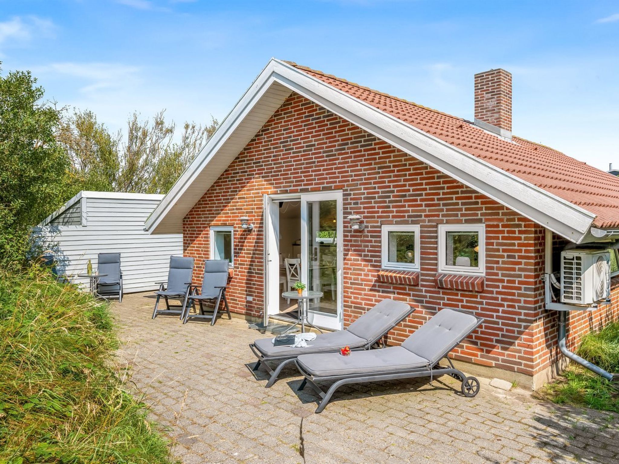 Une maison en briques avec un toit en pente, une terrasse avec deux chaises longues et une petite table.