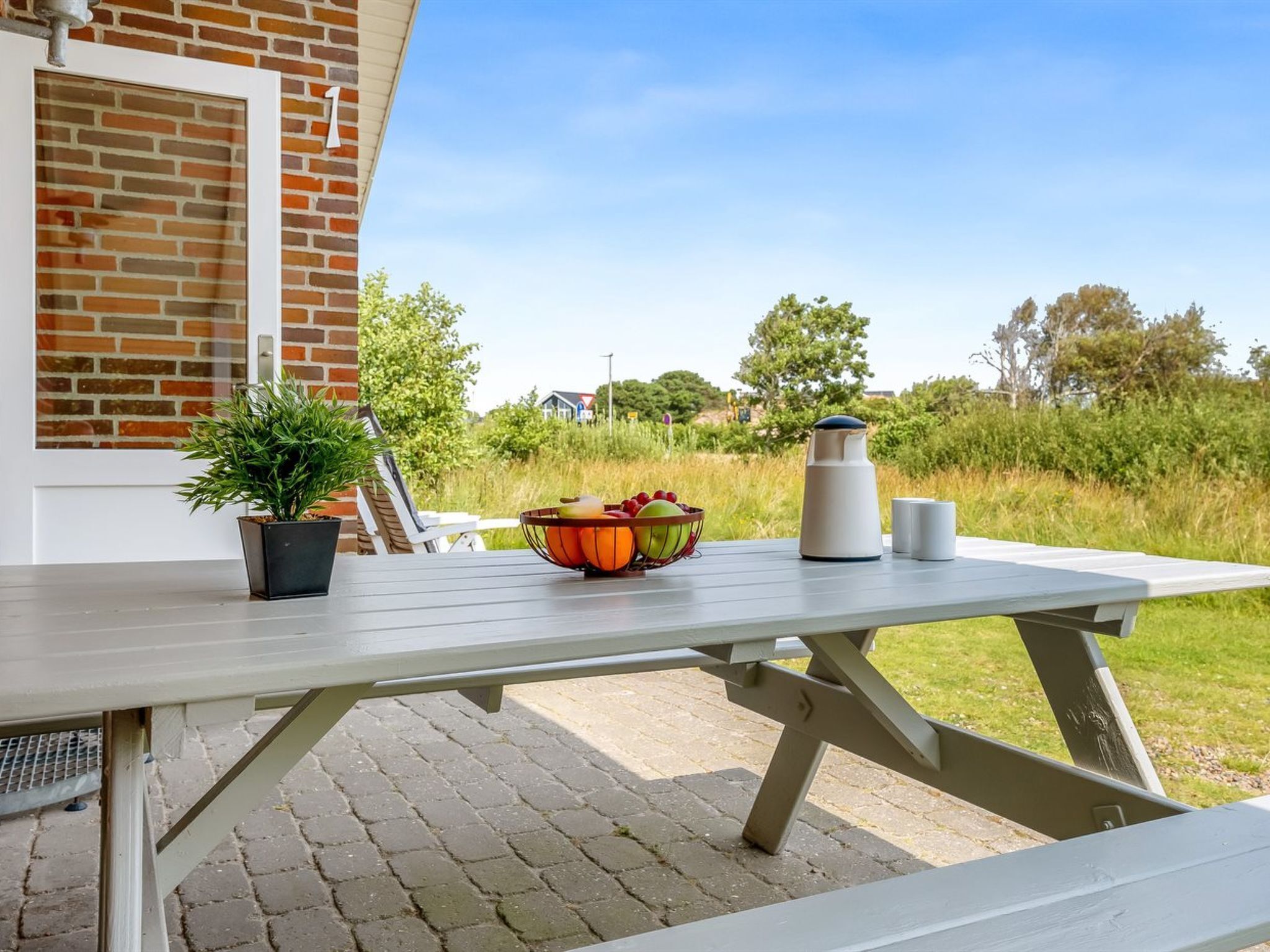 A picnic table with a basket of fruit, a kettle, and a cup outside a brick building.