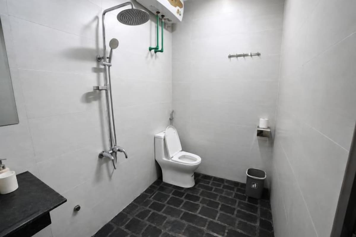 A bathroom with a white toilet, a shower with a handheld showerhead, a trash bin, and a towel rack.