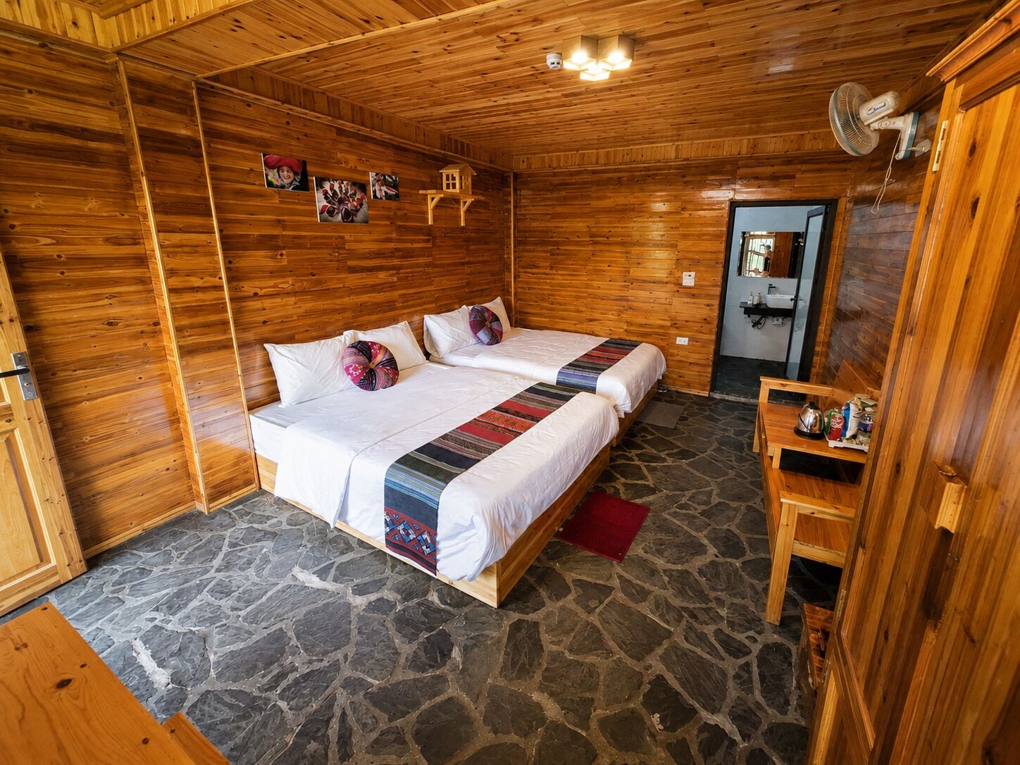 A wooden cabin room with two beds, a stone floor, a wooden door, and a small table with items on it.