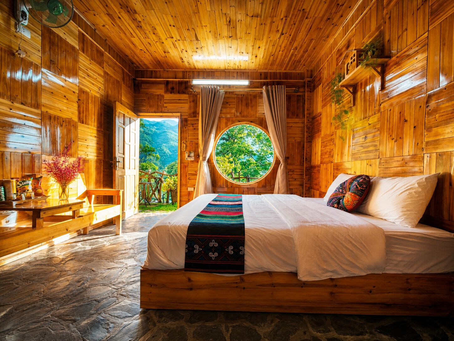 Habitación de cabaña de madera con cama, una mesa con aperitivos, un ventilador y ventana con vista a follaje.