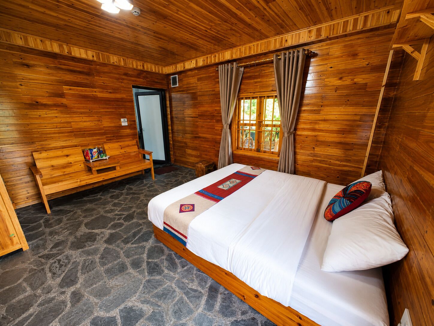 A wooden-paneled room with a bed, a bench, and a window with curtains.