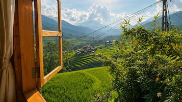 Un cadre de fenêtre en bois offrant une vue sur un paysage verdoyant, comprenant des champs en terrasses et une chaîne de montagnes.