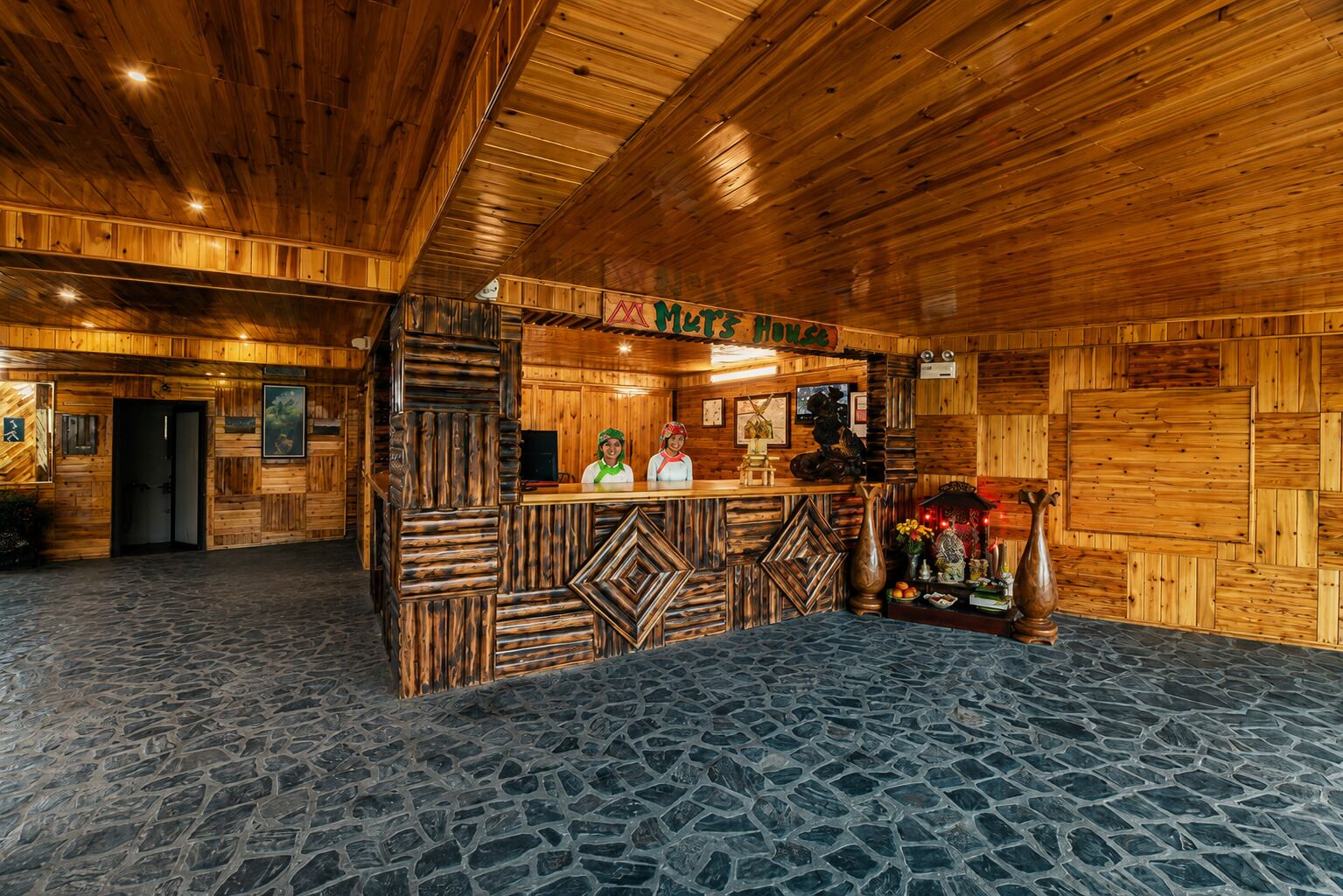 A wooden interior with a reception desk, stone flooring, and a sign that reads 'MUTZ HOUSE'.