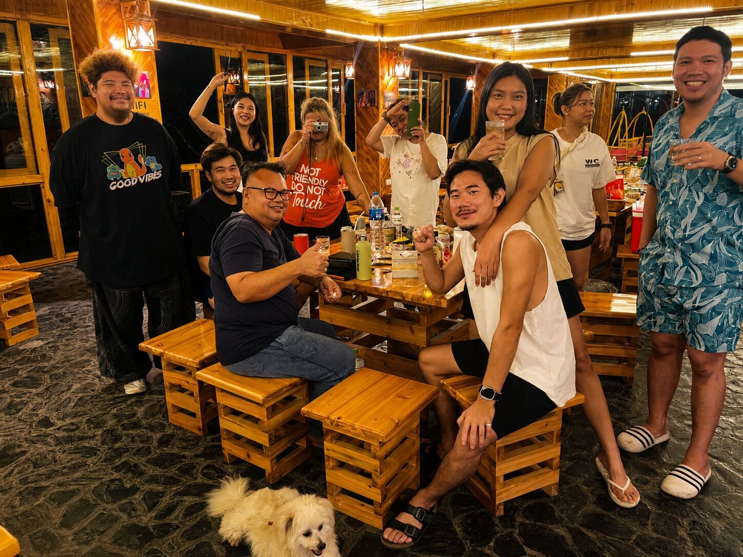 A group of people seated at wooden tables in a room with a stone floor, wooden walls, and hanging lights.