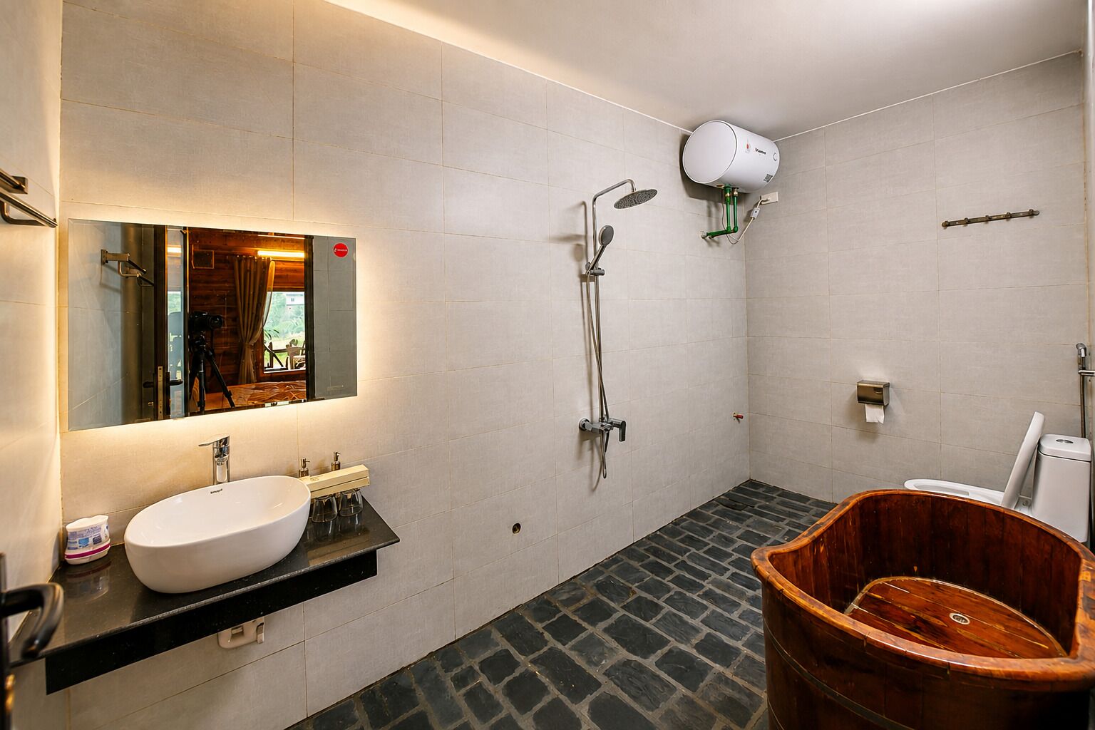 Un baño moderno con un espejo grande, un lavamanos blanco, una ducha con cabezal de ducha manual y un cubo de madera.