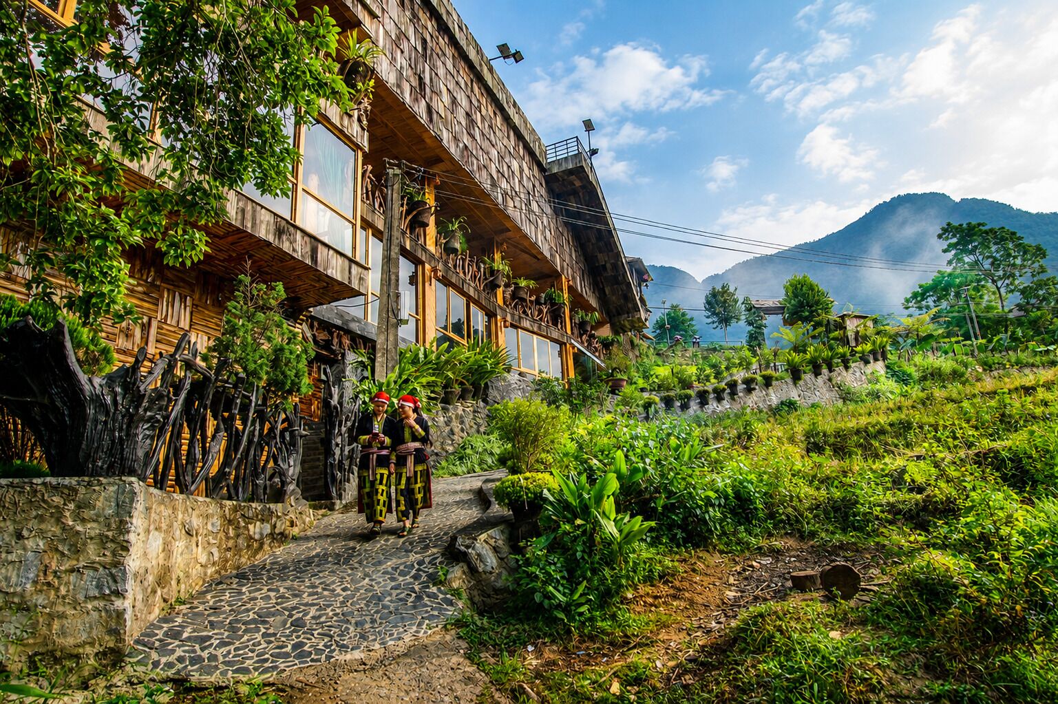 A traditional wooden building with a stone pathway leading to it, surrounded by lush greenery and a mountain in the background.