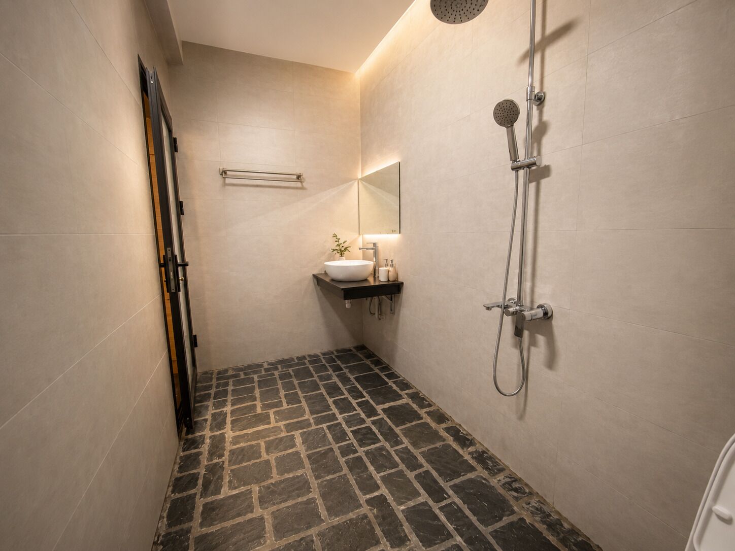 Un baño moderno con piso de baldosas de piedra, ducha con rociador circular y lavamanos con espejo.