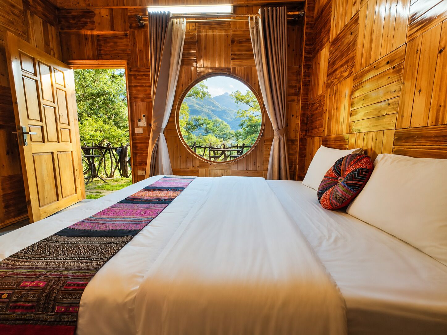 Una habitación de madera con una cama, un ventanal con vistas a zonas verdes y una puerta.