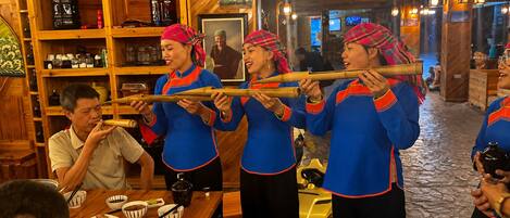 Un grupo de personas con vestimenta tradicional tocando un instrumento musical en un interior de madera con estantes y una mesa con comida.