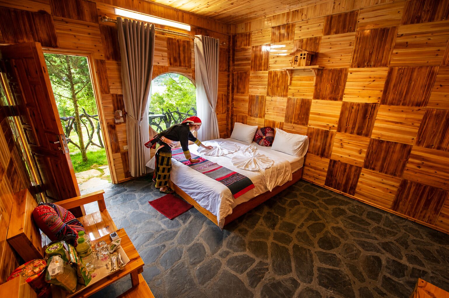 A wooden cabin room with a bed, a table with items, and a view of greenery outside.