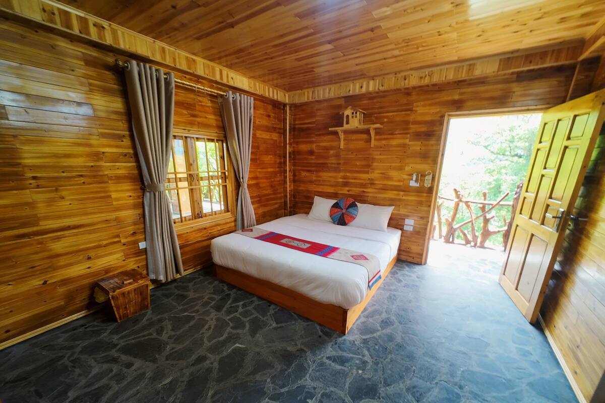 Ein Schlafzimmer einer Holzhütte mit Steinboden, einem Bett mit roter und blauer Decke und einem Fenster mit Vorhängen.