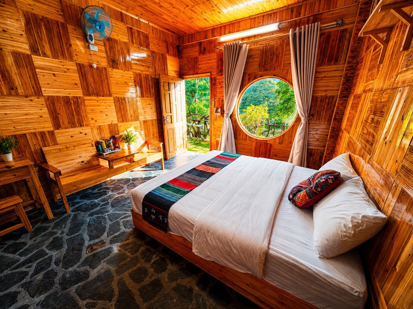 A wooden room with a bed, a round window, a stone floor, and a view of greenery outside.