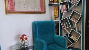 Una poltrona color petrolio, un tavolino in vetro con un vaso di fiori, un calendario da parete e una libreria con libri e oggetti decorativi.