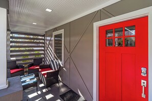 Een overdekt terras met rieten meubels en rode kussens, een rode deur met een keypad slot, en uitzicht op de straat door een raam.