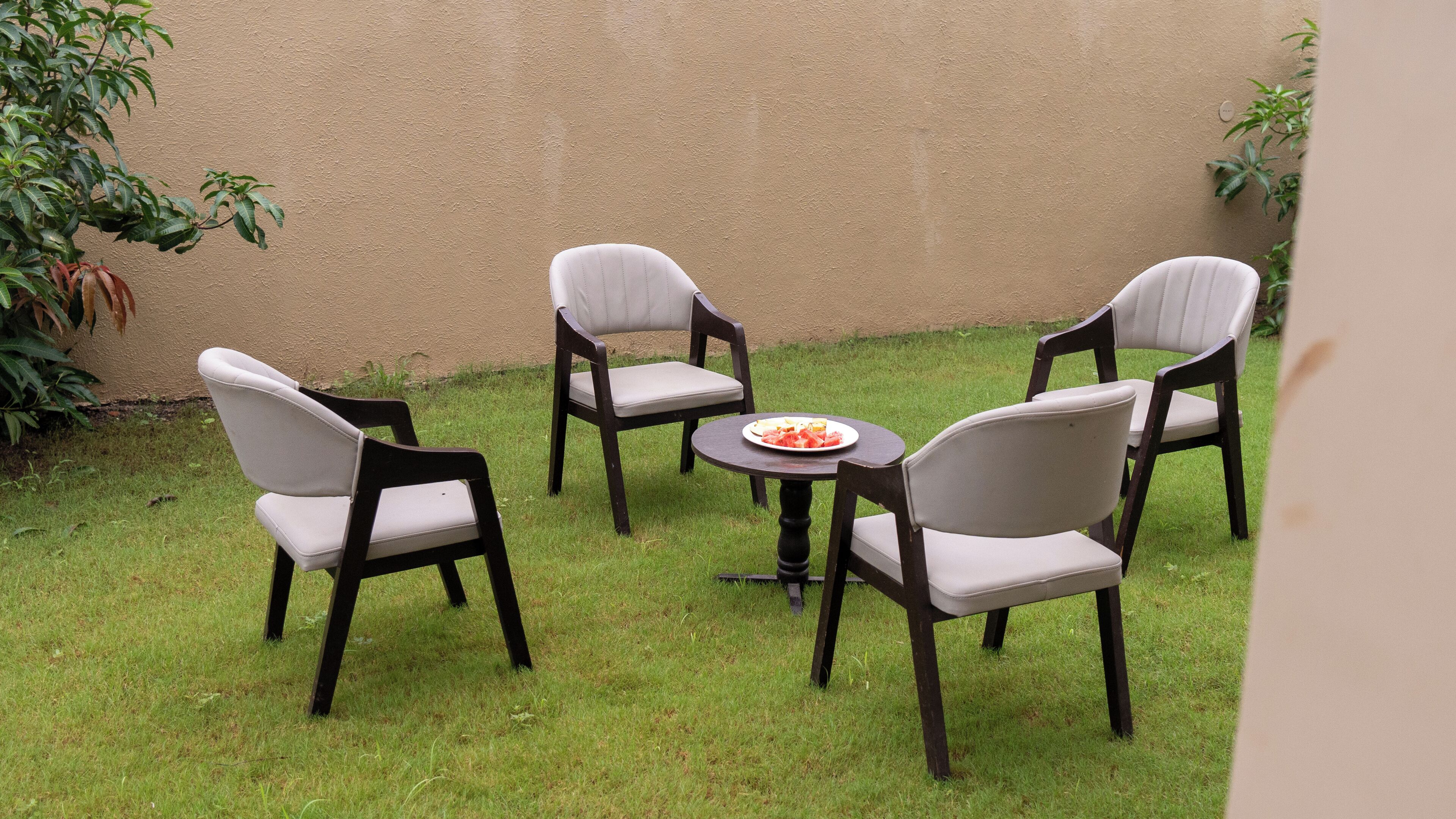 A small outdoor seating area with four chairs and a table, surrounded by a grassy lawn and a beige wall.