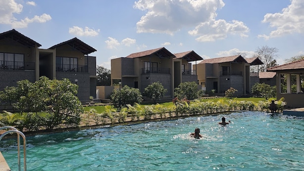 A swimming pool surrounded by modern buildings with balconies, a clear sky, and lush greenery. - The premier gir resort (Talala)