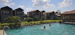 A swimming pool surrounded by modern buildings with balconies, a clear sky, and lush greenery. - The premier gir resort (Talala)