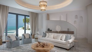 Un salon moderne doté d’une grande fenêtre donnant sur la plage, d’une table basse ronde en bois et d’un canapé blanc agrémenté de coussins gris.