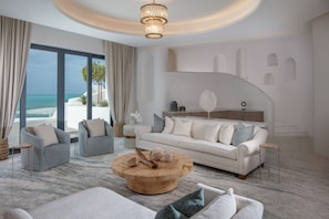 Una sala de estar moderna con un amplio ventanal con vista a la playa, una mesa de centro redonda de madera y un sofá blanco con cojines grises.
