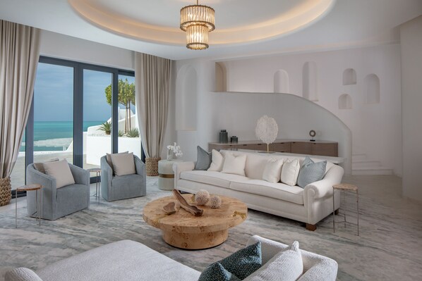 Una sala de estar moderna con un amplio ventanal con vista a la playa, una mesa de centro redonda de madera y un sofá blanco con cojines grises.