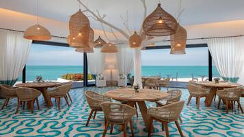 A seaside restaurant with round tables and wicker chairs, large hanging wicker lamps, and a view of the ocean.