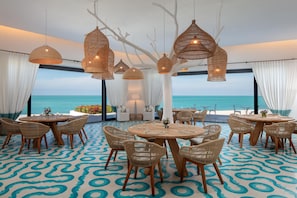 Un restaurant en bord de mer avec des tables rondes et des chaises en osier, de grandes lampes en osier suspendues et une vue sur l’océan.
