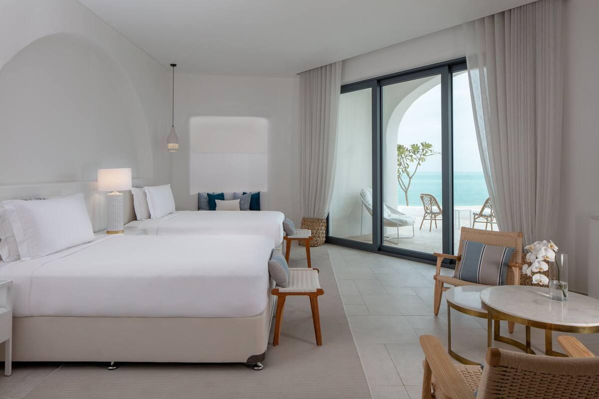Una habitación de hotel moderna con una cama grande, un área de estar con una mesa y sillas, y un balcón con vista al mar.