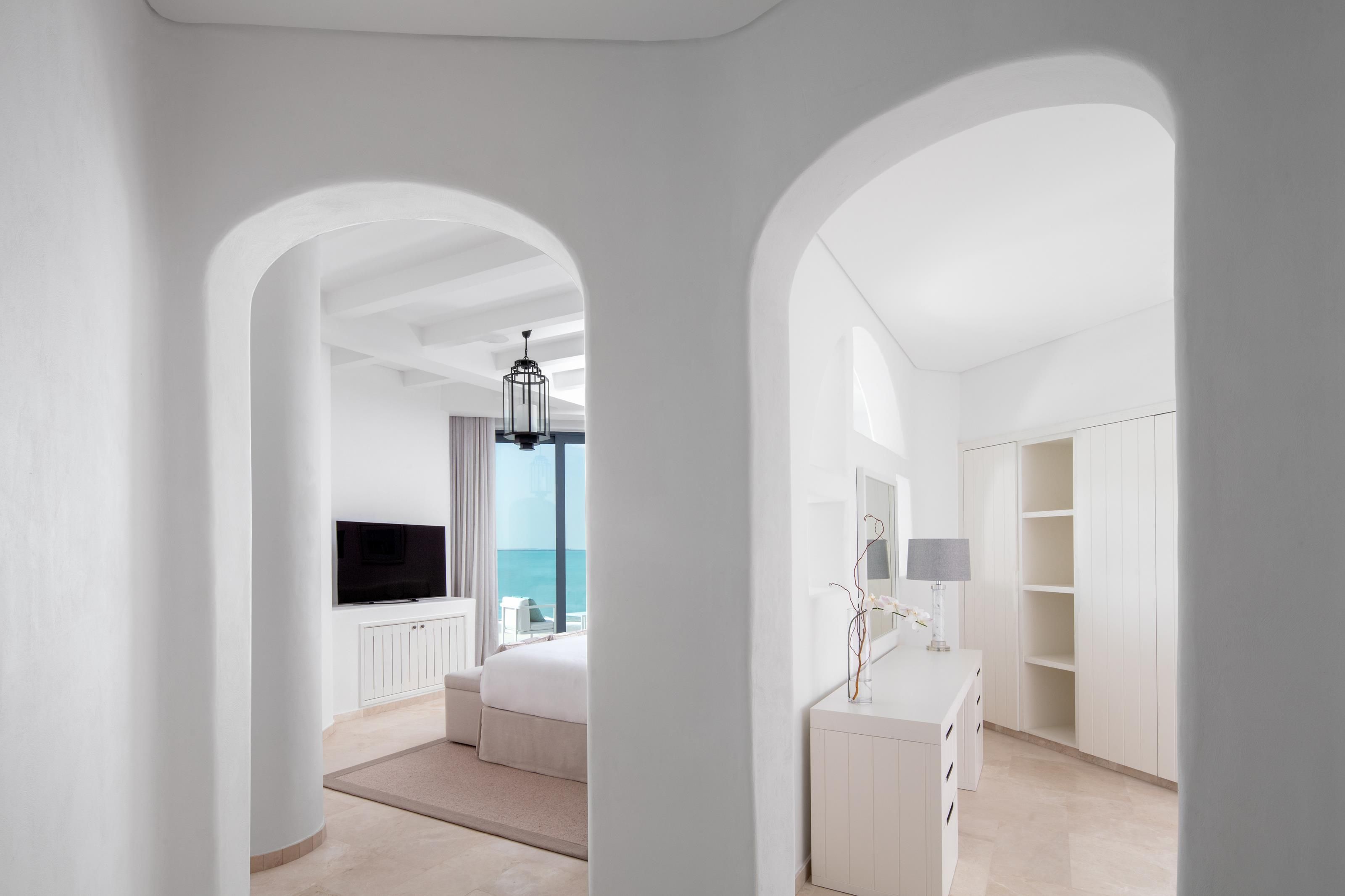A modern hotel room with a large bed, a TV, a desk with a lamp, and a view of the sea through a window.