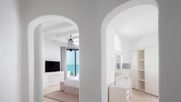 A modern hotel room with a large bed, a TV, a desk with a lamp, and a view of the sea through a window.