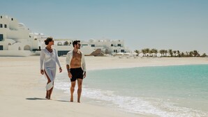 Two people walking on a beach with white buildings in the background. - Anantara Retreat Santorini Abu Dhabi - Adults Only (Ghantoot)