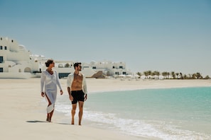 Deux personnes se promènent sur une plage, avec des bâtiments blancs en arrière-plan.