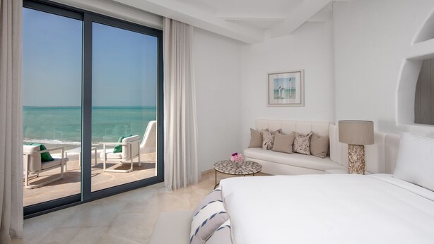 A hotel room with a large sliding glass door leading to a balcony with a view of the ocean. There is a bed, a sofa, a small round table with a lamp, and a framed picture on the wall. - Anantara Retreat Santorini Abu Dhabi - Adults Only (Ghantoot)