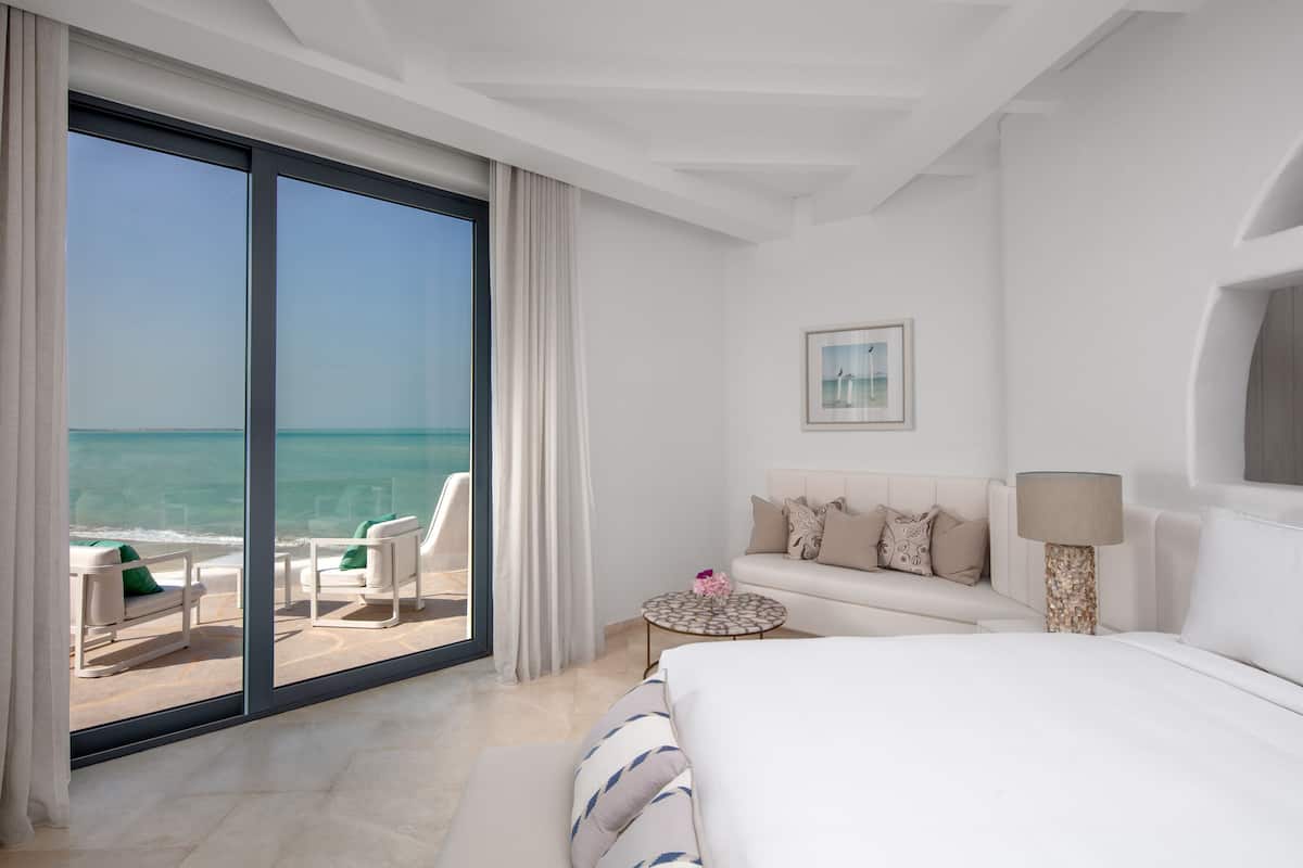 Habitación de hotel con un amplio ventanal corredero que da a un balcón con vista al océano. Se observa una cama, un sofá, una pequeña mesa redonda con una lámpara y un cuadro en la pared.