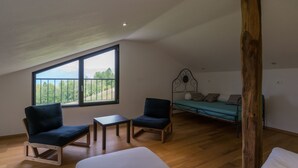 Une pièce avec un plafond en pente, une grande fenêtre donnant sur un paysage verdoyant, un lit, deux chaises et une petite table.