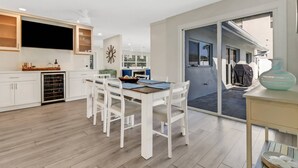 A modern kitchen with white cabinetry, a dining area with white chairs, and a large window overlooking a patio. - 5609 Del Rio Ct (Cape Coral)