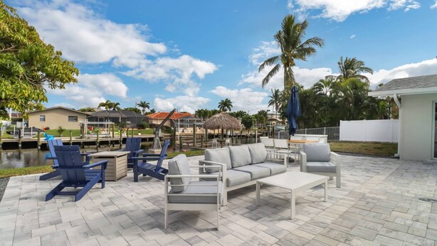 An outdoor patio with a sofa, chairs, and a table. There are palm trees and a body of water in the background. - 5609 Del Rio Ct (Cape Coral)