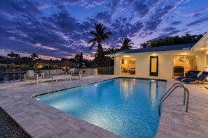 A swimming pool with lounge chairs, a covered seating area, and a building with a door and windows. - 5609 Del Rio Ct (Cape Coral)