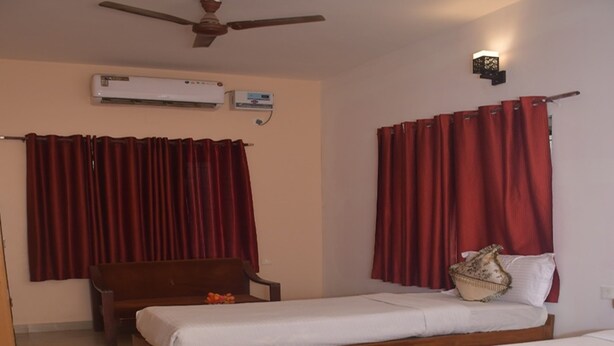 Habitación de hotel con dos camas individuales, una silla de madera, ventilador de techo, aire acondicionado y cortinas rojas.