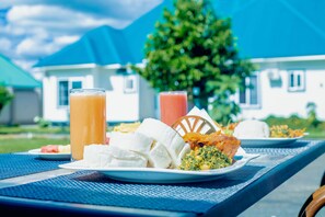 A dining table with a meal consisting of rice, vegetables, and a piece of meat, set against a backdrop of a house and a tree. - Teranga Lodge & Restaurant (Mitumba)