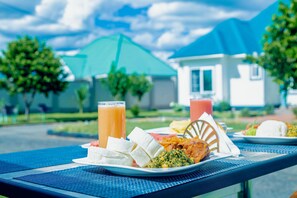 A dining table with plates of food and drinks, a chair, and a blue umbrella in a courtyard with buildings in the background. - Teranga Lodge & Restaurant (Mitumba)