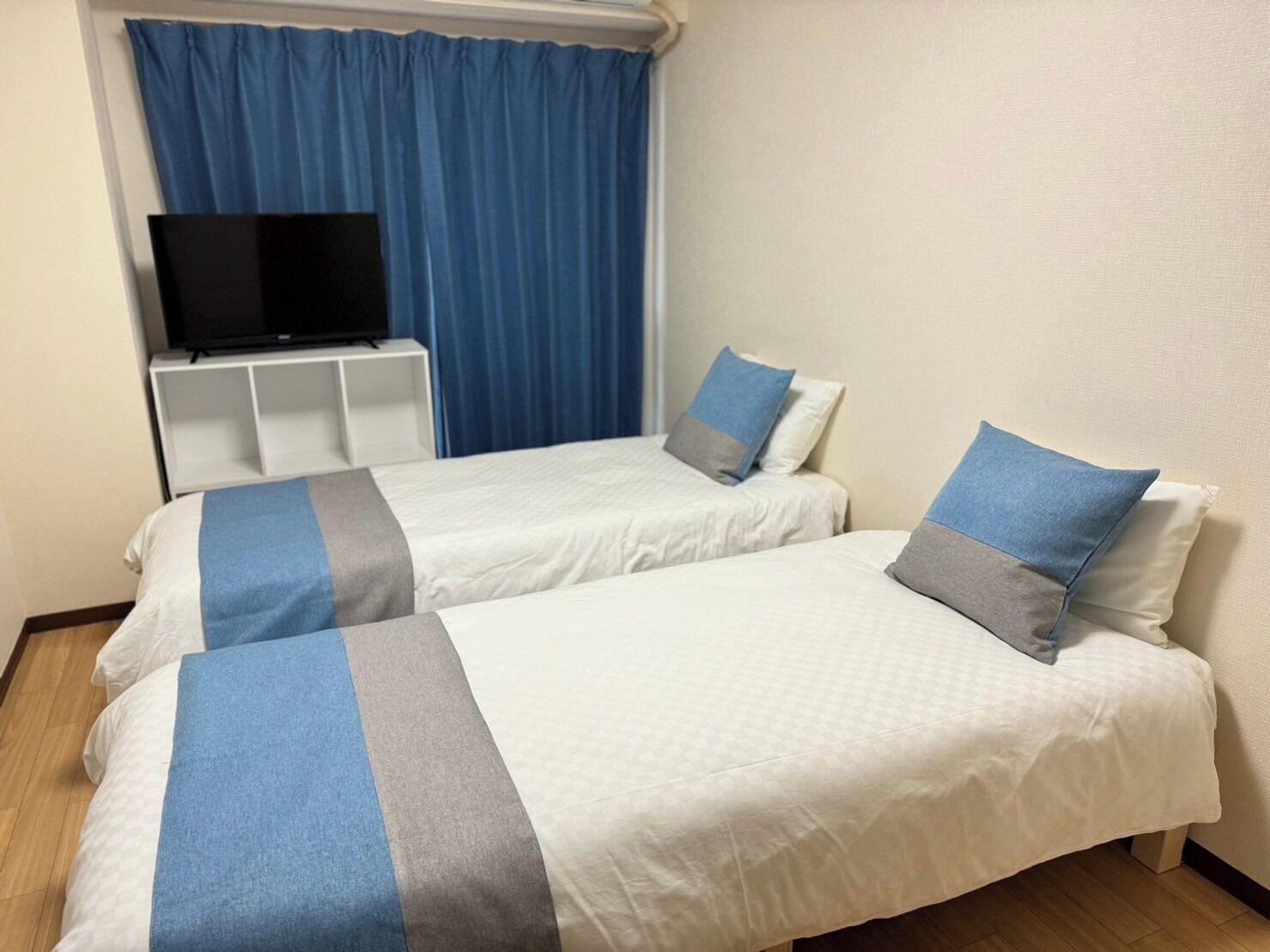 A hotel room with two beds, a TV on a stand, and a window with blue curtains.
