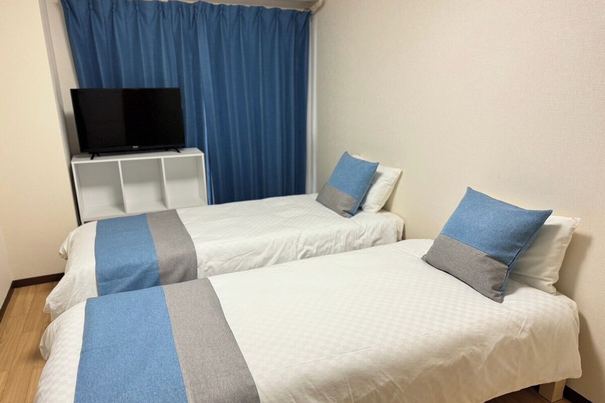 Ein Hotelzimmer mit zwei Betten, einem Fernseher auf einem Tisch und einem Fenster mit blauen Vorhängen.
